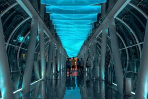People in the middle of a a futuristic hallway lit by a bright blue ceiling.