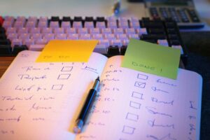 Hand written to do list in a notebook with a fountain pen lying on the book. A yellow post-it-note stuck to the book with handwritten text that says 'to do' and a green post-it-note with a handwritten text that says "done". A pink and black computer keyboard in the background out of focus.