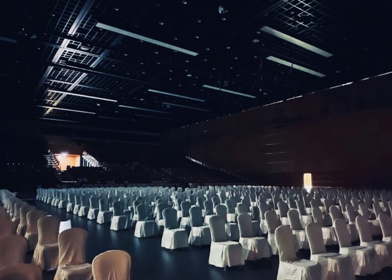Photo of dark conference room taken from the stage looking across rows of chairs covered in white fabric seat covers with the entrance at the rear lit prior to the crowd entering.