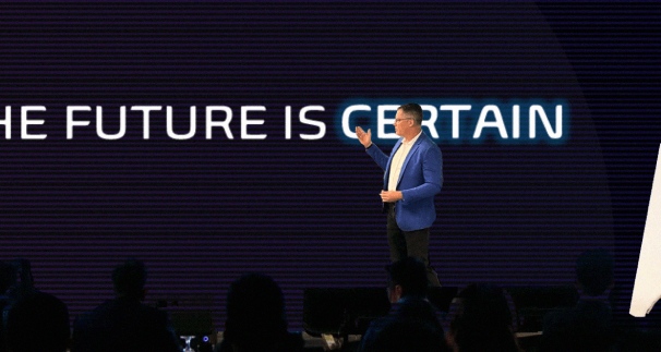 Brett Wiskar delivering keynote to audience in front of large screen displaying white text that reads "The Future if Certain" 
