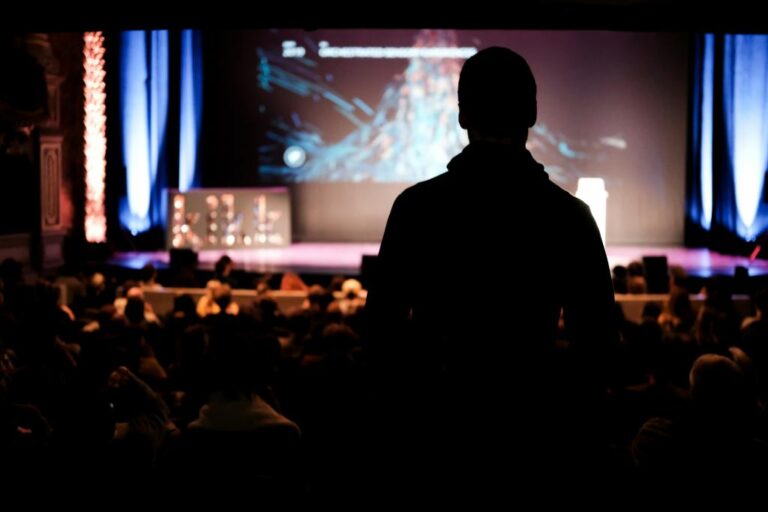 A silhouette of an AV technician in foreground looking on to out of focus conference stage and audience. Photo by Cosmin Serban, Unsplash