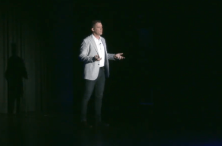 Brett Wiskar delivering a keynote presentation from the stage Brett Wiskar delivering a keynote presentation from the stage. Large image of Brett in isolation on a dark stage. He's wearing a grey jacket and gesturing to the audience.