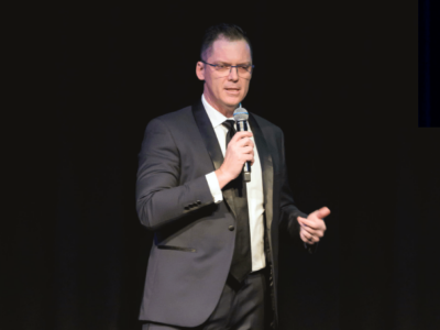 Brett Wiskar delivering a keynote to a formal event Brett Wiskar delivering a keynote to a formal event in a dinner suit using a handheld microphone.