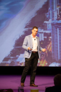 Brett Wiskar Speaking to a large franchisee audience at Royal Pines on the Gold Coast Brett Wiskar Speaking to a large franchisee audience at Royal Pines on the Gold Coast with a large screen behind him showing a photo of the nearby beach and buildings.